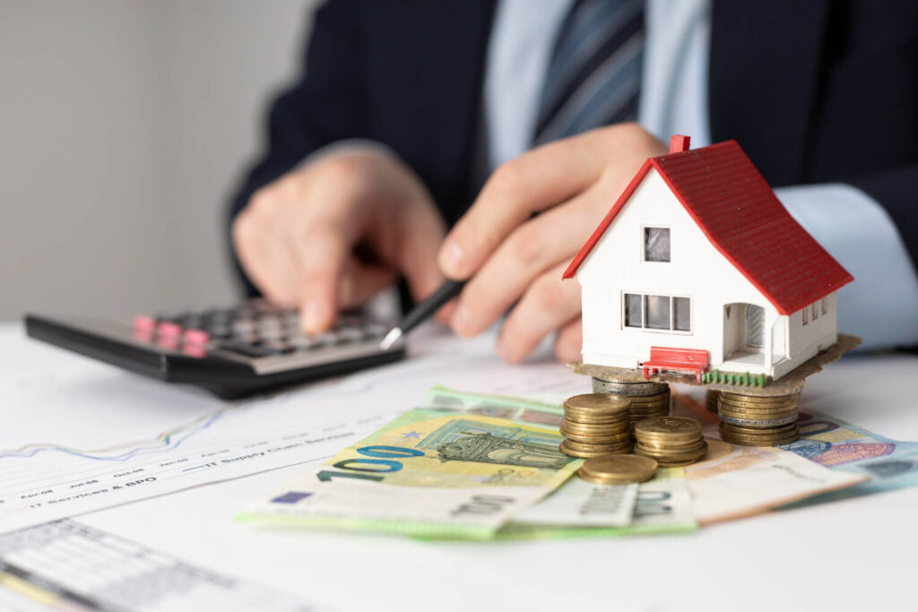 Das Bild zeigt im Vordergrund ein Modellhaus mit rotem Dach, das auf mehreren gestapelten Euromünzen steht. Darunter liegen verschiedene Eurobanknoten, hauptsächlich 100- und 50-Euro-Scheine. Im Hintergrund ist eine Person im Anzug und mit Krawatte zu sehen, die mit einem Kugelschreiber eine Taste auf einem Taschenrechner drückt. Auf dem Tisch liegen zudem ein Blatt mit Diagrammen und eine Tabelle. Die Szene ist scharf auf das Hausmodell und das Geld im Vordergrund fokussiert, während der Hintergrund leicht verschwommen ist.