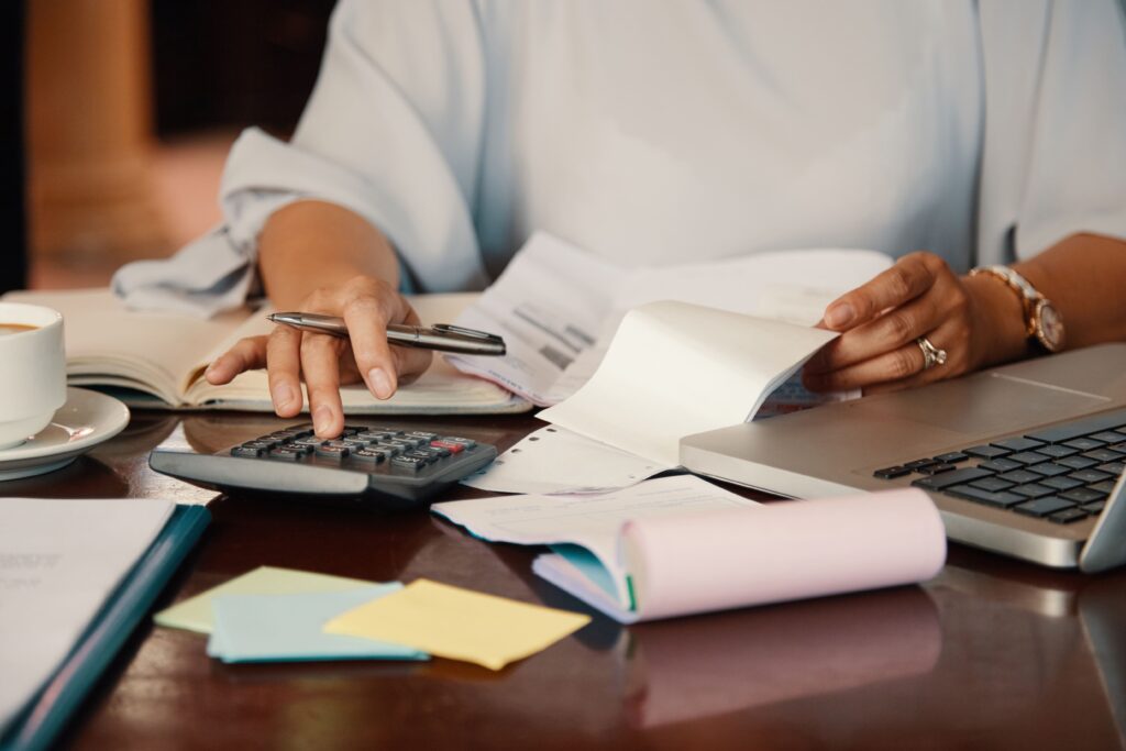 Nahaufnahme einer Person, die an einem Tisch arbeitet. Die Person trägt eine hellblaue Bluse und ist nur von den Schultern abwärts sichtbar. Sie hält einen Stift in der rechten Hand und tippt mit der linken auf einen Taschenrechner. Auf dem Tisch liegen ein geöffneter Laptop, verschiedene Dokumente, ein Notizbuch, ein Kassenblock, farbige Haftnotizen und eine Tasse Kaffee auf einer Untertasse. Die Person trägt eine Armbanduhr und einen Ring an der linken Hand. Die Szene vermittelt eine Arbeitssituation mit Fokus auf Buchhaltung oder Finanzen.