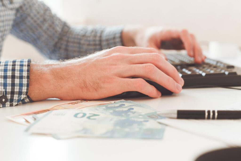 Ein Mann mit kariertem Hemd tippt an einer Tastatur. Auf dem Tisch liegen mehrere Euro-Geldscheine und ein Kugelschreiber. Die Szene ist nah aufgenommen.